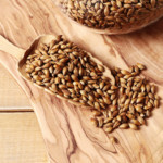 Graines d&rsquo;orge grill&eacute;s dispos&eacute;s dans une cuilli&egrave;re en bois, qui d&eacute;borde sur son support, une planche en bois.