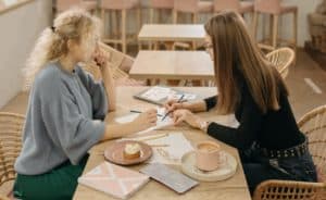Deux filles blanches (une blonde, un brune) travaillent autour d&rsquo;une table carr&eacute;e en bois, dans un restaurant 