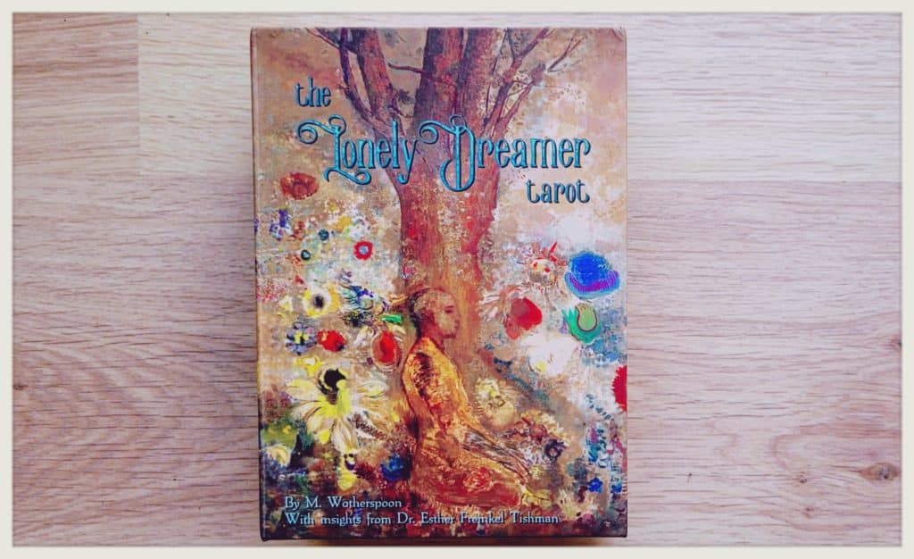 La boite du Lonely Dreamer tarot sur du parquet couleur naturelle. L&rsquo;illustration est dans le style symboliste : sur un fond ocre se dessine un arbre presque esquiss&eacute; marron. Tout autour, des motifs de fleurs presques abstraites peintes &agrave; l&rsquo;huile en jaune, rouge, et bleu, font ressortir des ton bleut&eacute;s au niveau du sol, qui ne se distingue pas du ciel. On y devine, dans un style entre l&rsquo;impressionnisme et l&rsquo;abstrait, un m&eacute;ditant qui n&rsquo;a pas &eacute;t&eacute; peint &agrave; l&rsquo;exception de sa toge en jaune ocre.